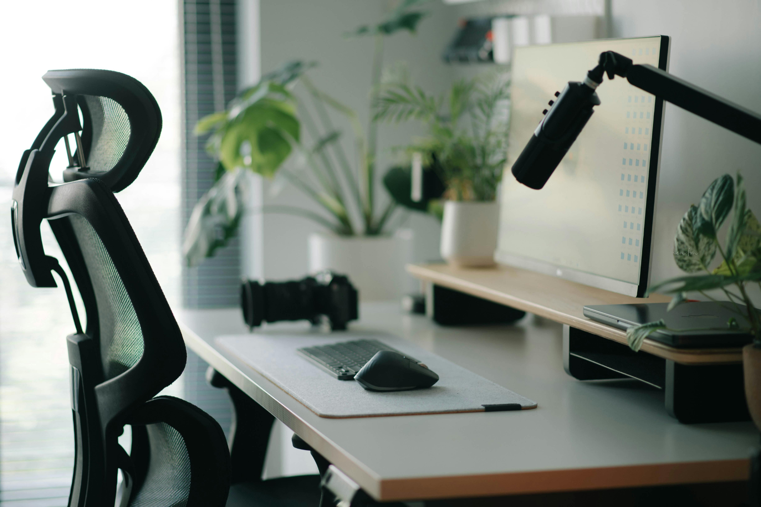 Modern home office desk setup with a black office chair and headrest, computer monitor, keyboard, microphone, and indoor plants